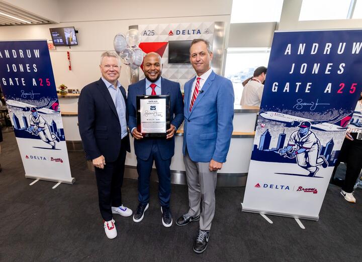 Three people pose at a Delta airport gate ceremony, with the center person holding a commemorative plaque. Banners on both sides read “Andruw Jones Gate A25,” featuring baseball-themed artwork and Delta branding.