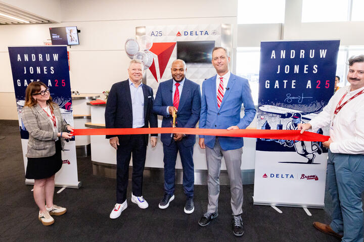Five people pose at a Delta airport gate ceremony, with the center person holding large scissors to cut a red ribbon. Banners on both sides read “Andruw Jones Gate A25,” featuring baseball-themed artwork and Delta branding.
