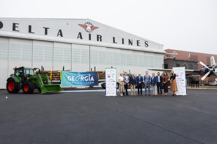 Group stands in front of Delta Air Lines’ historic hangar at the Delta Flight Museum alongside a tractor, timber load and Georgia SAF Coalition signage during a sustainable aviation fuel event.