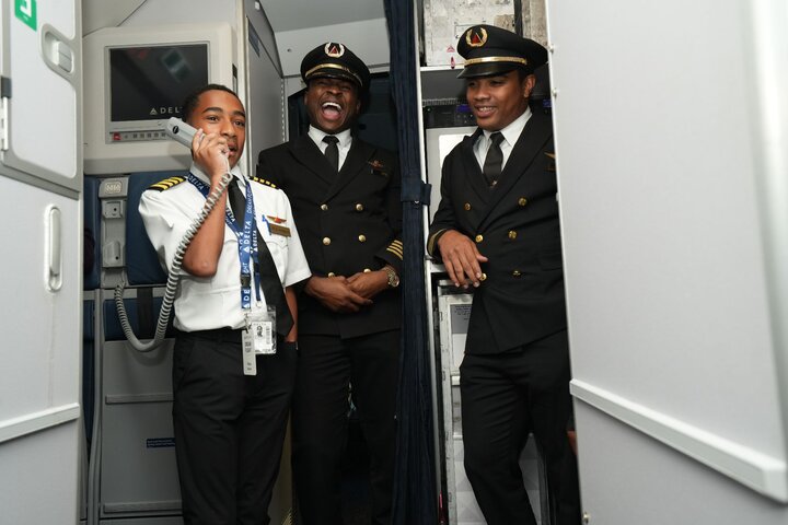 DREAM Flight participant makes an onboard announcement from the aircraft galley while Delta pilots look on.