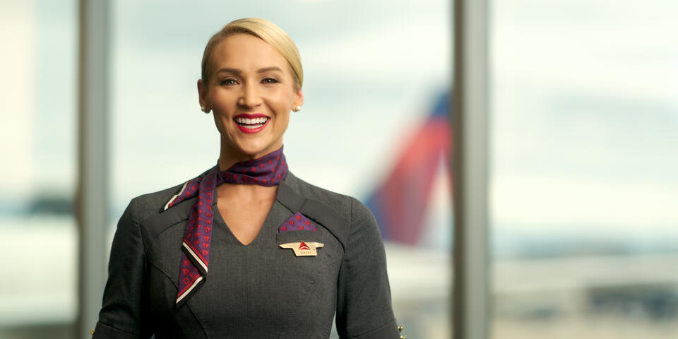 Delta flight attendant smiling, wearing gray dress and purple neck scarf.