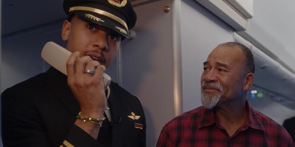 Airline pilot in uniform stands inside an aircraft cabin near the galley, holding an intercom handset. Another person wearing a red plaid shirt is standing beside the pilot.