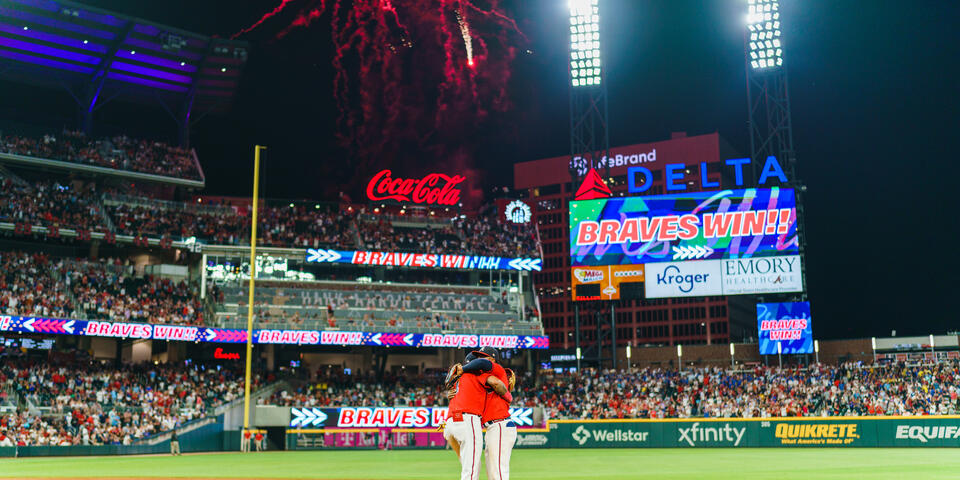 Fireworks light up a baseball stadium at night as two players stand together on the infield, with a scoreboard displaying “Braves Win” and a packed crowd in the background.