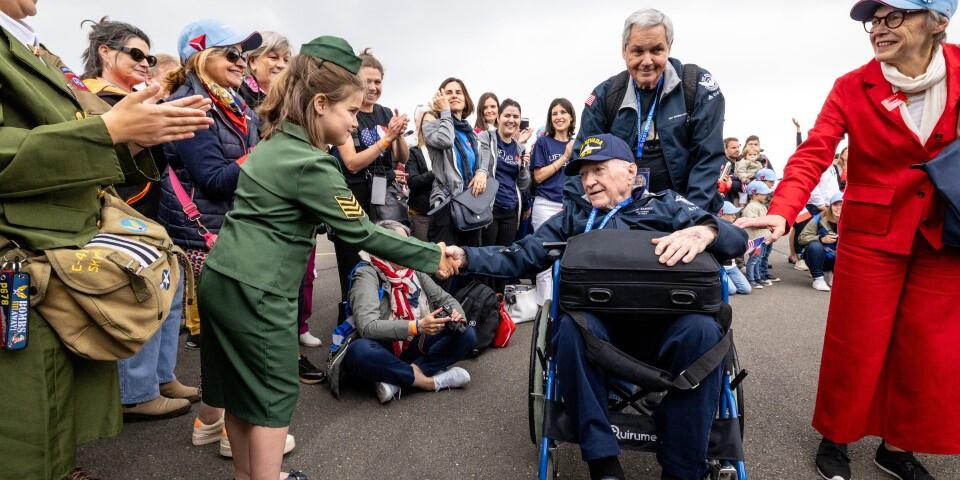 A seated veteran in a wheelchair shakes hands with a child in a green uniform as a crowd lines the roadway, applauding during a commemorative event.
