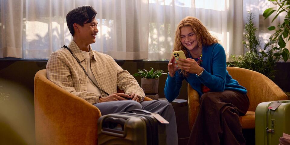 Two travelers sit in lounge chairs with rolling suitcases nearby, one looking at a smartphone, in a softly lit indoor waiting area with plants and sheer curtains.