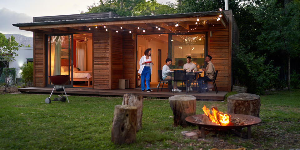 People dining outside a modern wooden cabin at dusk, with string lights, a fire pit and trees surrounding the yard.