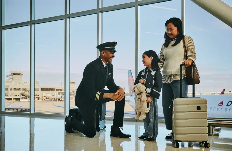 Delta pilot kneeling beside a young child holding a stuffed animal at an airport gate, while an adult stands nearby with rolling luggage; airplane visible through large windows in the background.