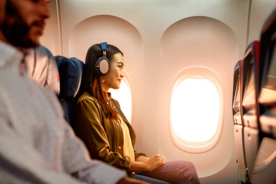 Delta customer on board an airplane wearing headphones sitting by a window, with warm sunlight illuminating the cabin and seatback screens visible.