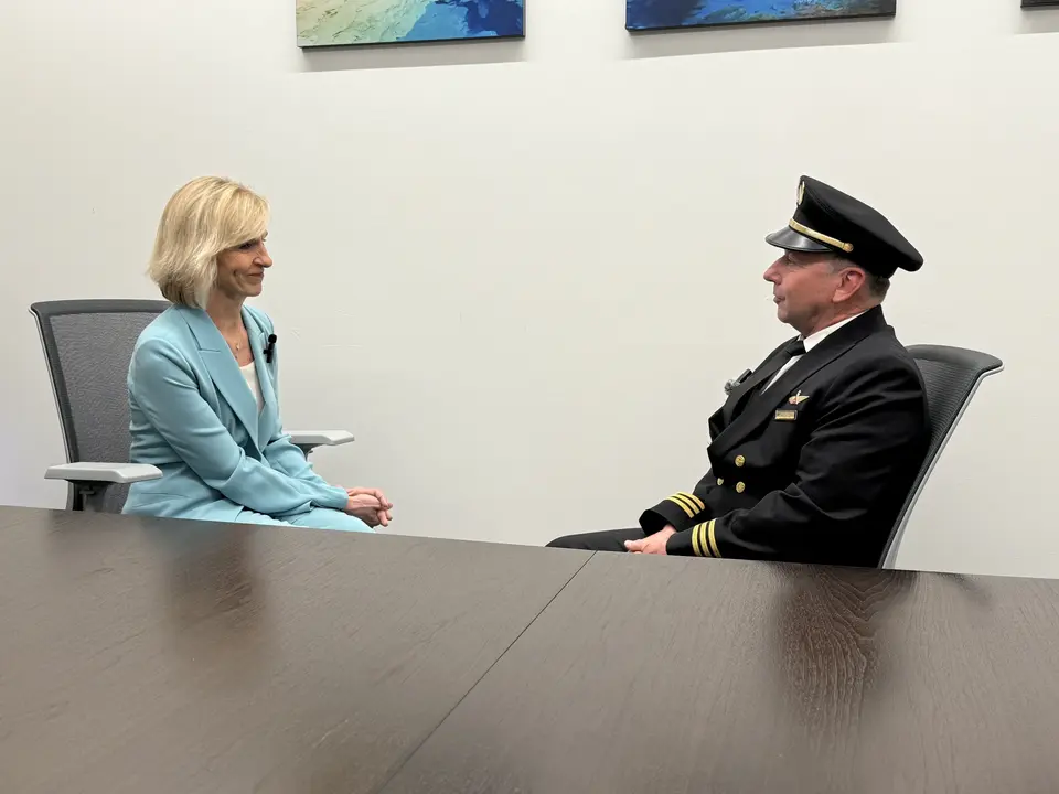 Allison Ausband and Tom Cohan sit facing each other at a conference table, Allison in a business suit and Tom in a pilot uniform.