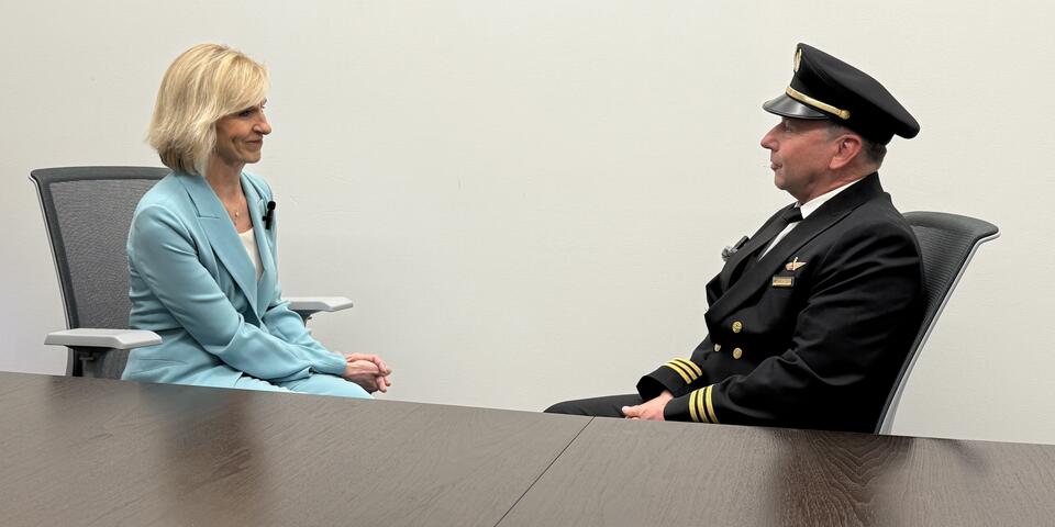 Allison Ausband and Tom Cohan sit facing each other at a conference table, Allison in a business suit and Tom in a pilot uniform.