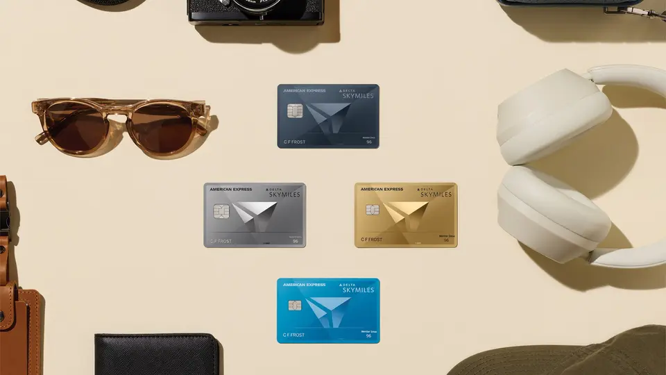 Flat lay of Delta SkyMiles American Express Cards surrounded by travel essentials, including a passport, camera, sunglasses, headphones, and luggage accessories.