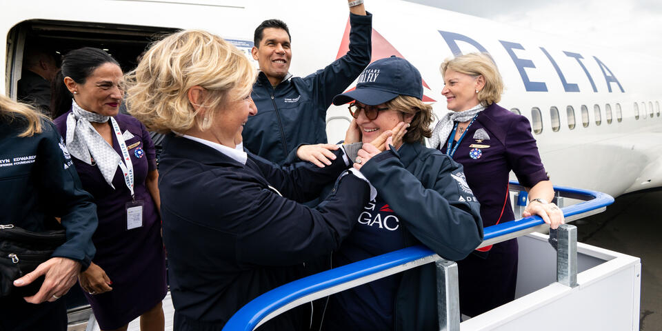 Delta employees and volunteers stand on an airport boarding ramp beside a Delta aircraft, welcoming a passenger as they exit the plane.