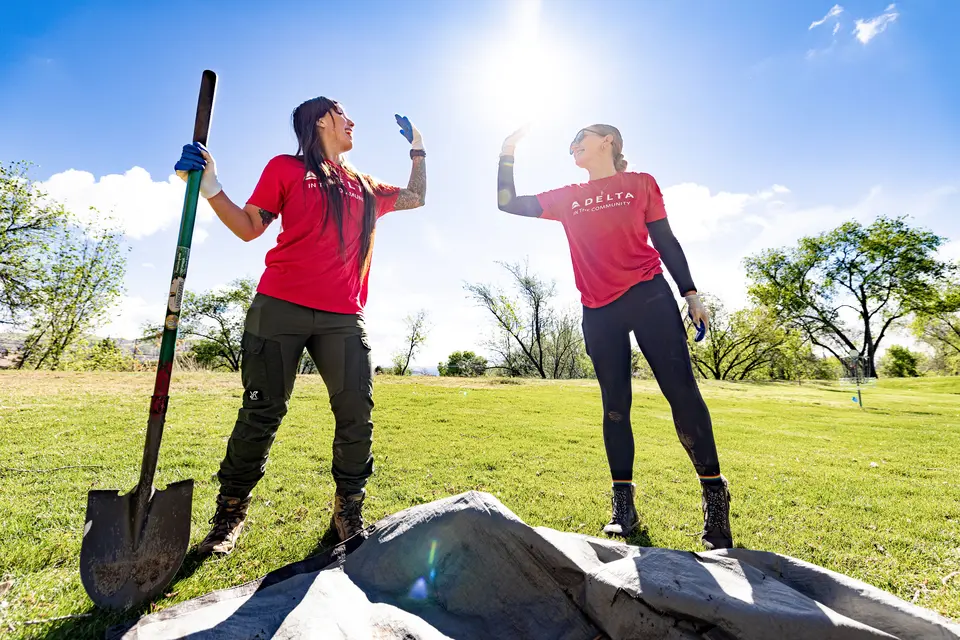Two volunteers in red “Delta in the Community” shirts high-five while working outdoors, one holding a shovel on a sunny grassy field with trees in the background.