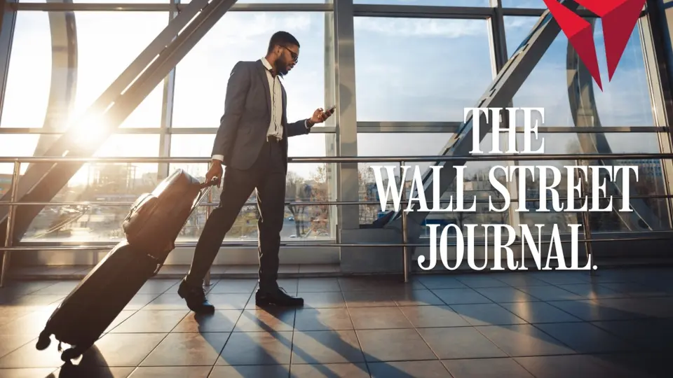 Traveler in a suit walks through a sunlit airport terminal, pulling a rolling suitcase and looking at a phone, with “The Wall Street Journal” text overlaid.