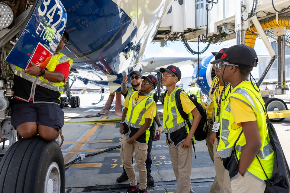 Students in safety vests gather beneath a Delta aircraft as an aviation professional points out features near the landing gear on the airport ramp.