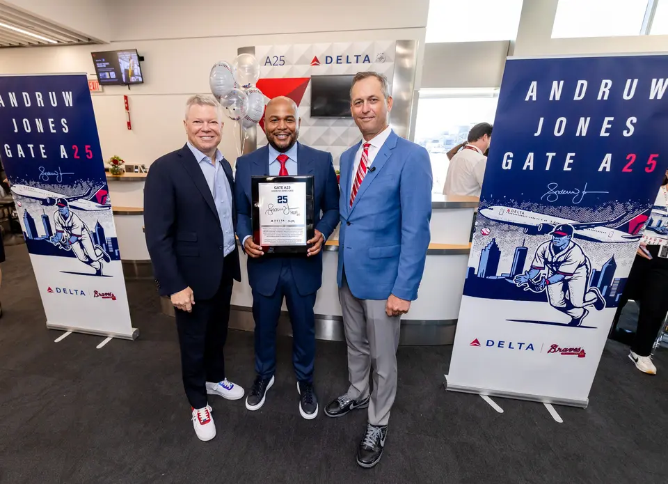 Three people pose at a Delta airport gate ceremony, with the center person holding a commemorative plaque. Banners on both sides read “Andruw Jones Gate A25,” featuring baseball-themed artwork and Delta branding.
