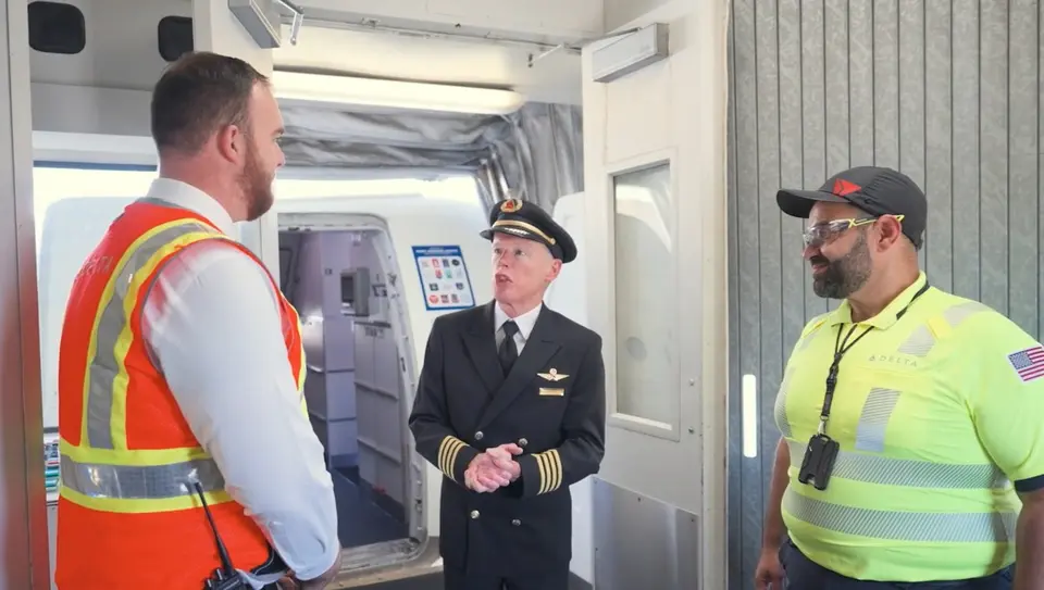 Three airport employees standing and talking near an aircraft boarding door, including a pilot in uniform and two workers wearing high-visibility safety vests.
