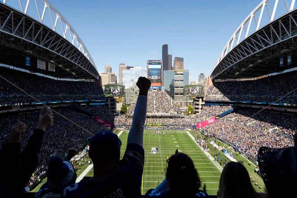 Fans in a packed football stadium watch the game from the stands, with the field and Seattle skyline visible beyond the open end of the venue.