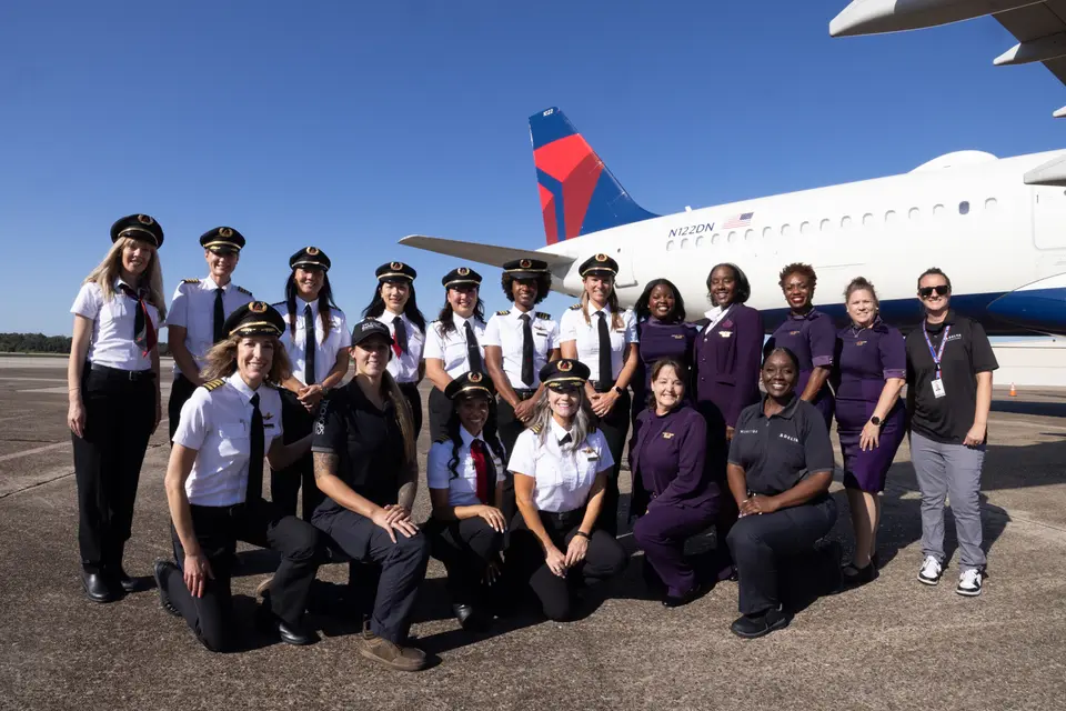 Group of aviation professionals and participants posing on the tarmac in front of a Delta aircraft under a clear blue sky during a Girls in Aviation Day event.