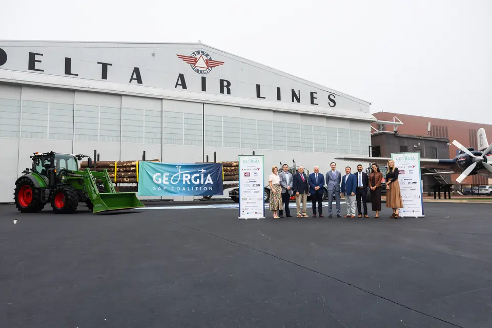 Group stands in front of Delta Air Lines’ historic hangar at the Delta Flight Museum alongside a tractor, timber load and Georgia SAF Coalition signage during a sustainable aviation fuel event.