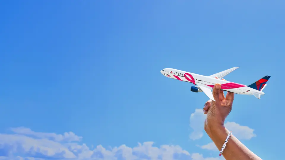 Hand holding a pink-branded Delta model airplane against a blue sky with clouds, symbolizing support for breast cancer awareness.