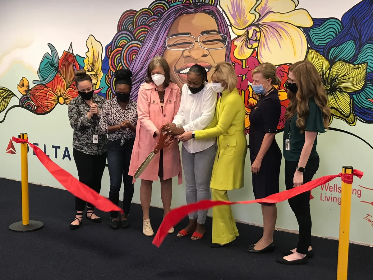 Wellspring and Delta leaders and employees cut a ribbon to present the final mural that symbolizes Delta's mission to combat human trafficking. The mural is of a smiling woman with flowers and rainbows behind her head. Beneath the image is the word 'free' and an imagery for Delta and Wellspring's partnership.