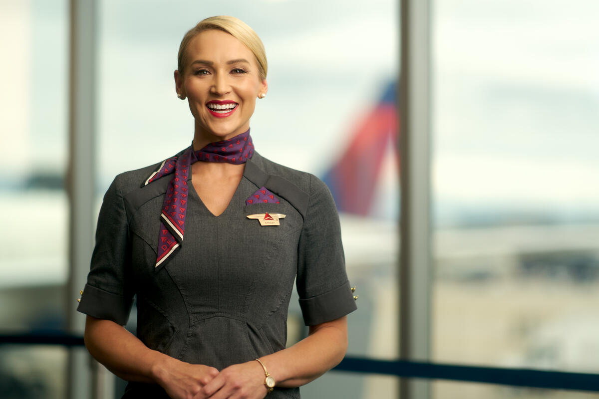 Delta flight attendant smiling, wearing gray dress and purple neck scarf.