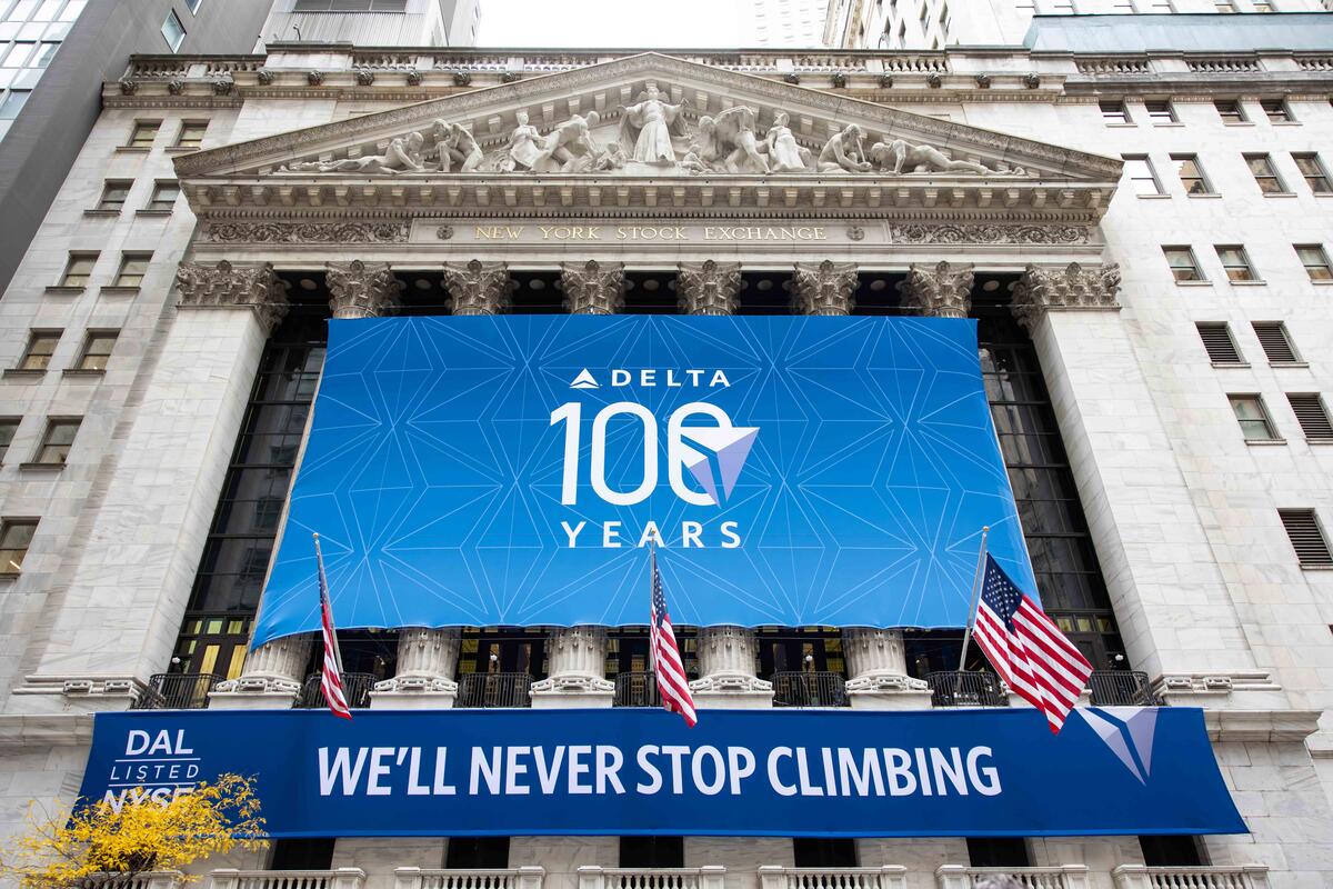Exterior shot of NYSE building with a large, rectangular banner displaying Delta's centennial logo and a smaller banner below that reads We'll Never Stop Climbing