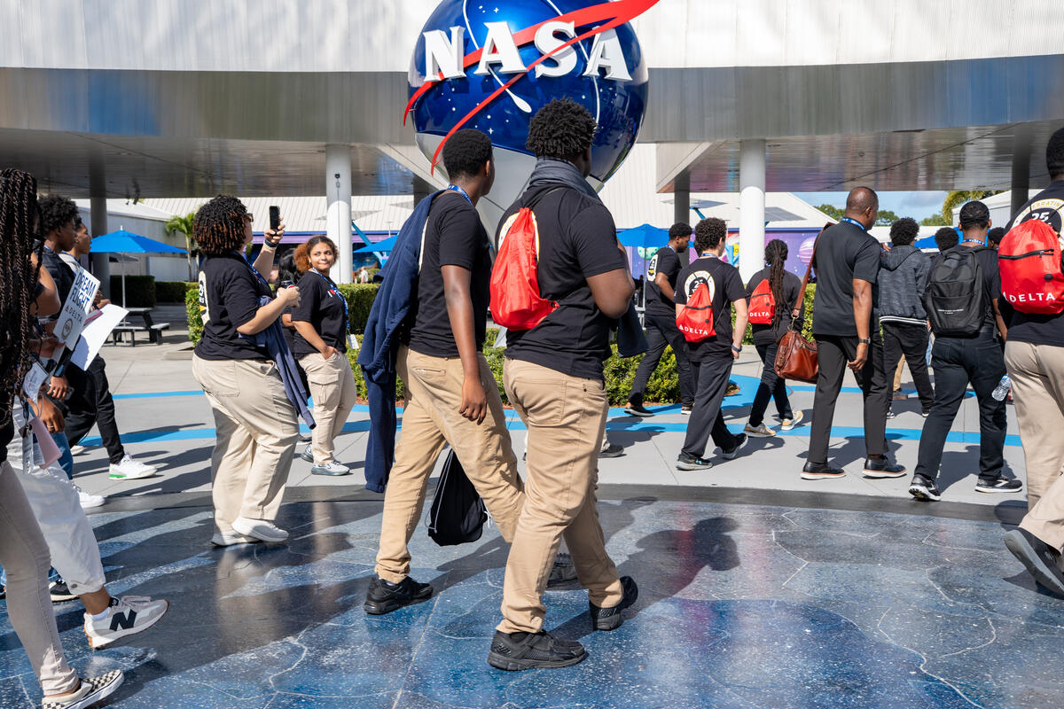DREAM flight participants walking near a large NASA logo sculpture outdoors at the Kennedy Space Center.