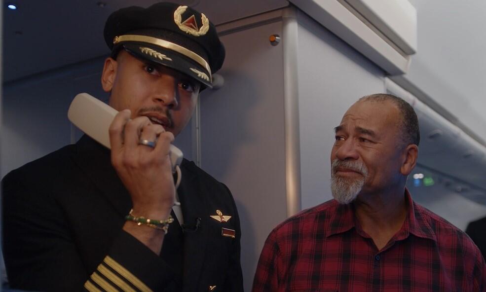 Airline pilot in uniform stands inside an aircraft cabin near the galley, holding an intercom handset. Another person wearing a red plaid shirt is standing beside the pilot.