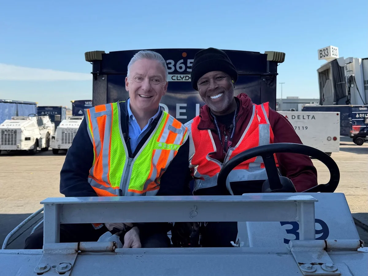 Two workers in high-visibility vests sitting on an airport ground vehicle near Delta equipment and a boarding gate.