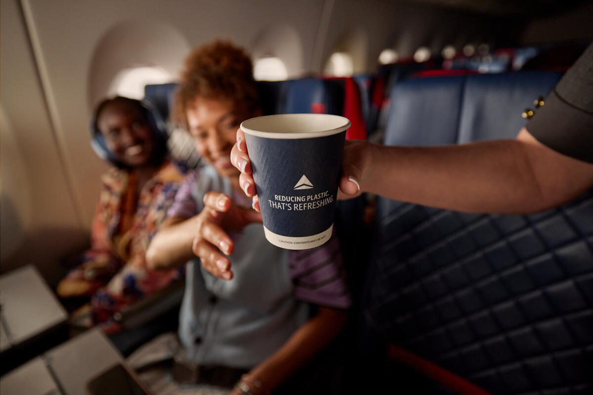 Delta's new paper cup in flight attendant's hand, ready to be handed to customer who is seated on board an aircraft and reaching for the cup.