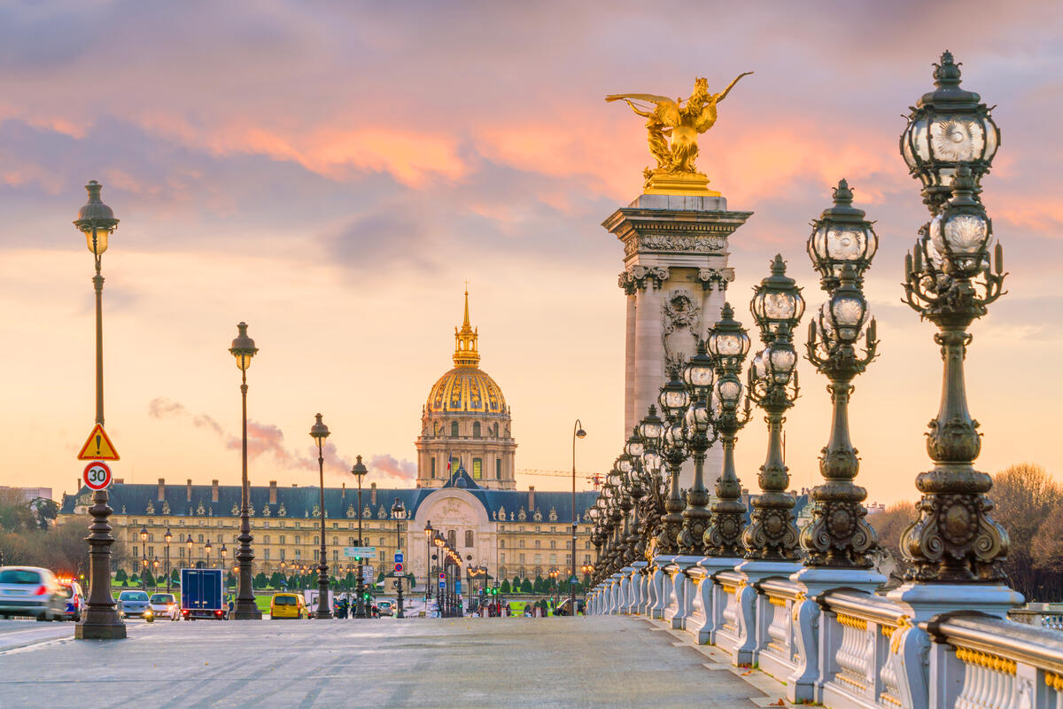 Ornate bridge lined with lamps at sunset, facing a domed historic building in the distance.