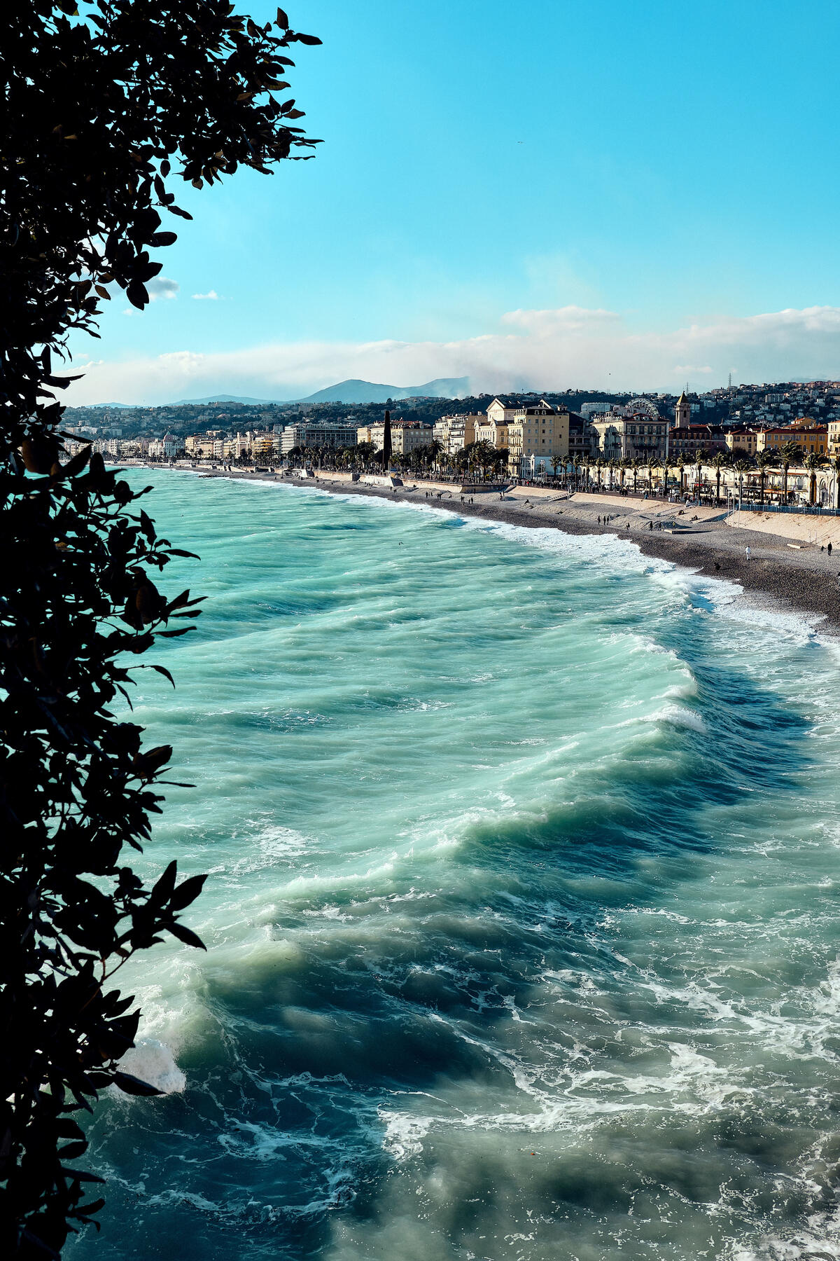 Waves rolling toward a long beachfront lined with buildings under a bright blue sky.