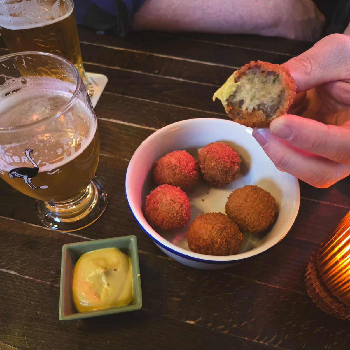 A bowl of crispy bitterballen with mustard, alongside a glass of beer and a hand holding a bitten bitterbal.