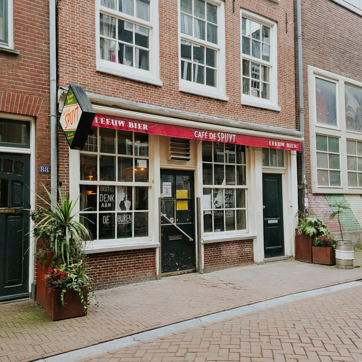 A narrow brick-front café with a red awning reading ‘Café de Spuyt,’ featuring large front windows, potted plants, and a quiet cobblestone street.