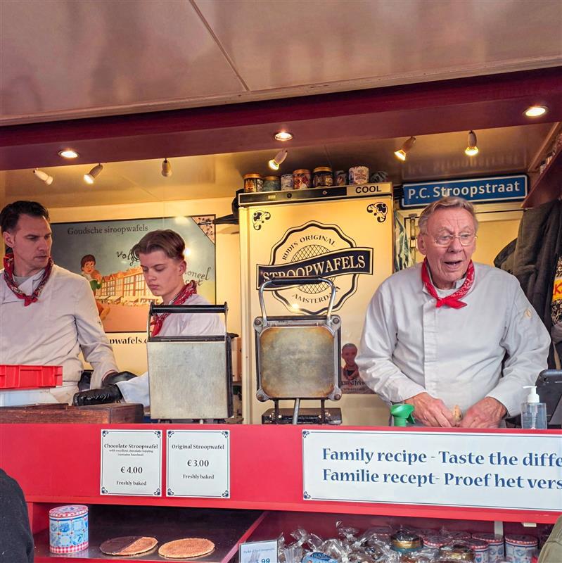 A stroopwafel stand with staff preparing fresh stroopwafels behind waffle irons, surrounded by signs and jars of ingredients.