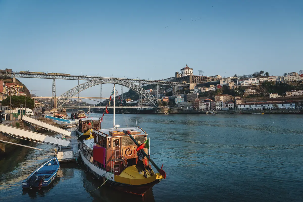 Colorful boats on a river with a large metal arch bridge and hillside buildings in the background.