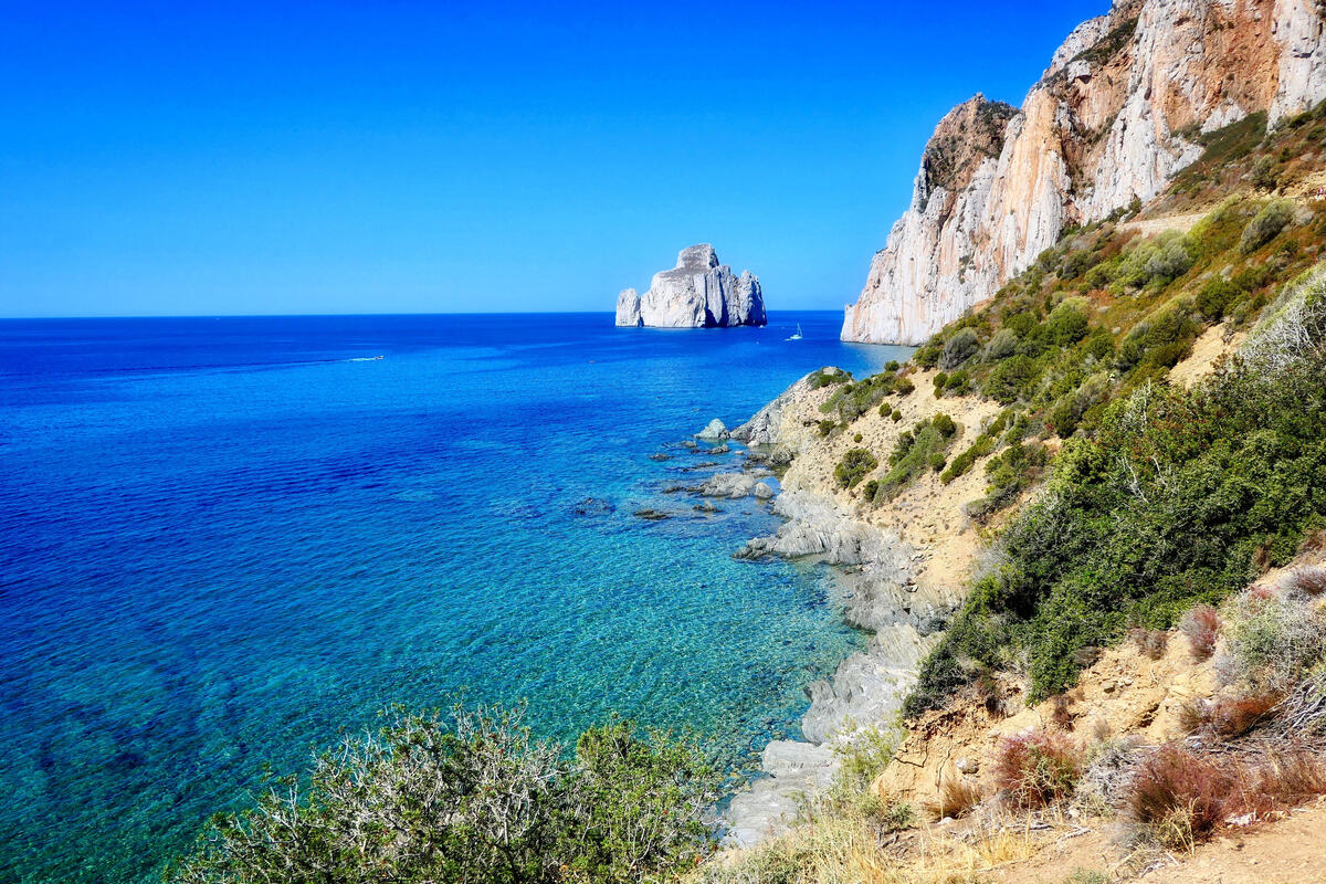 Clear turquoise sea beside rugged cliffs with a large rock formation offshore.