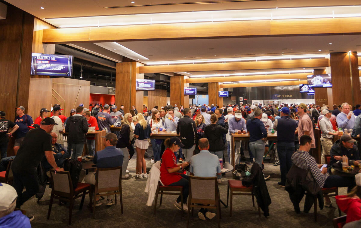 A crowded indoor stadium lounge with fans standing and sitting at tables beneath wood-paneled ceilings and multiple TV screens.