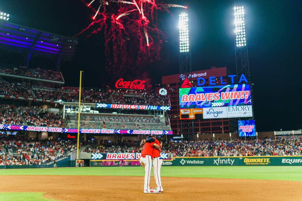 Fireworks light up a baseball stadium at night as two players stand together on the infield, with a scoreboard displaying “Braves Win” and a packed crowd in the background.
