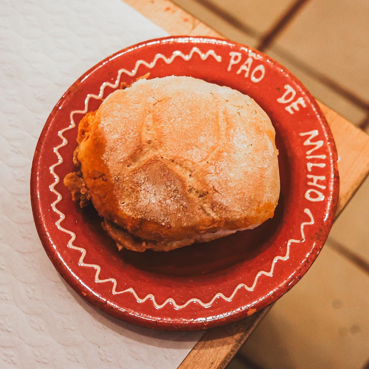 Portuguese bifana sandwich on a red ceramic plate labeled ‘Pão de Milho.’