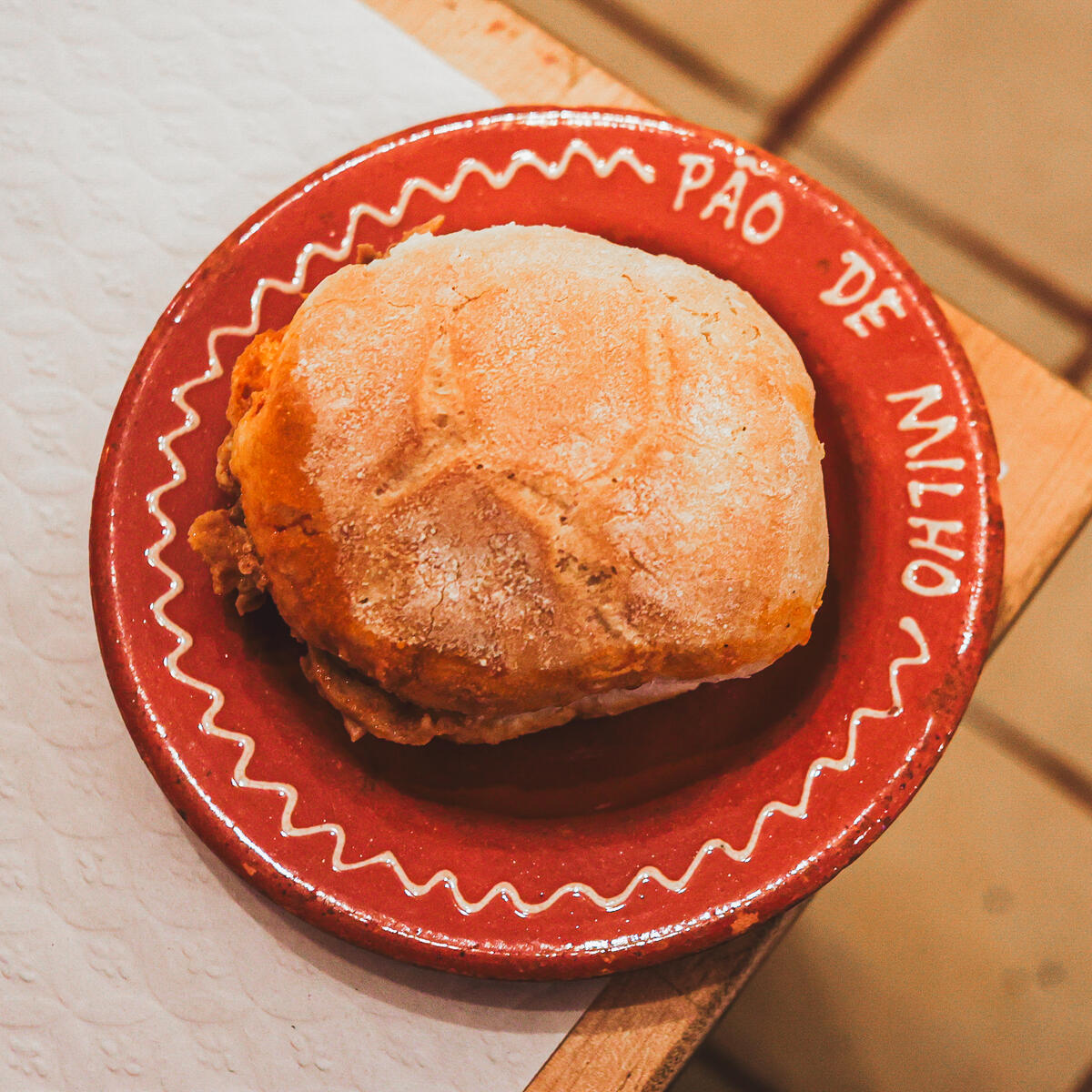 Portuguese bifana sandwich on a red ceramic plate labeled ‘Pão de Milho.’