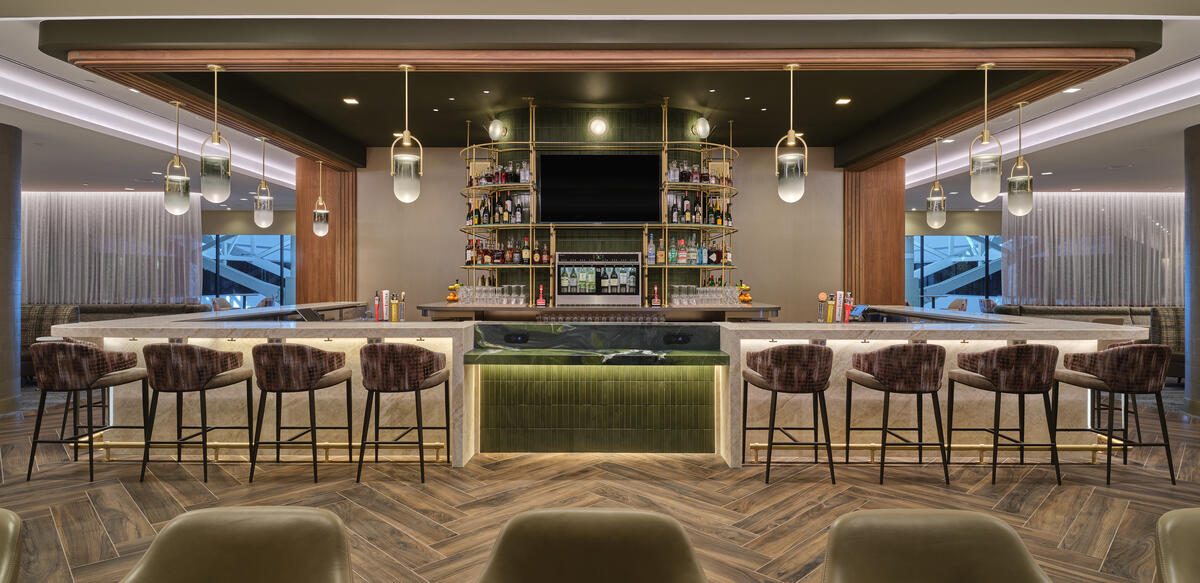 Front view of lounge bar with high‑back stools, illuminated shelving, pendant lights and wood‑pattern flooring