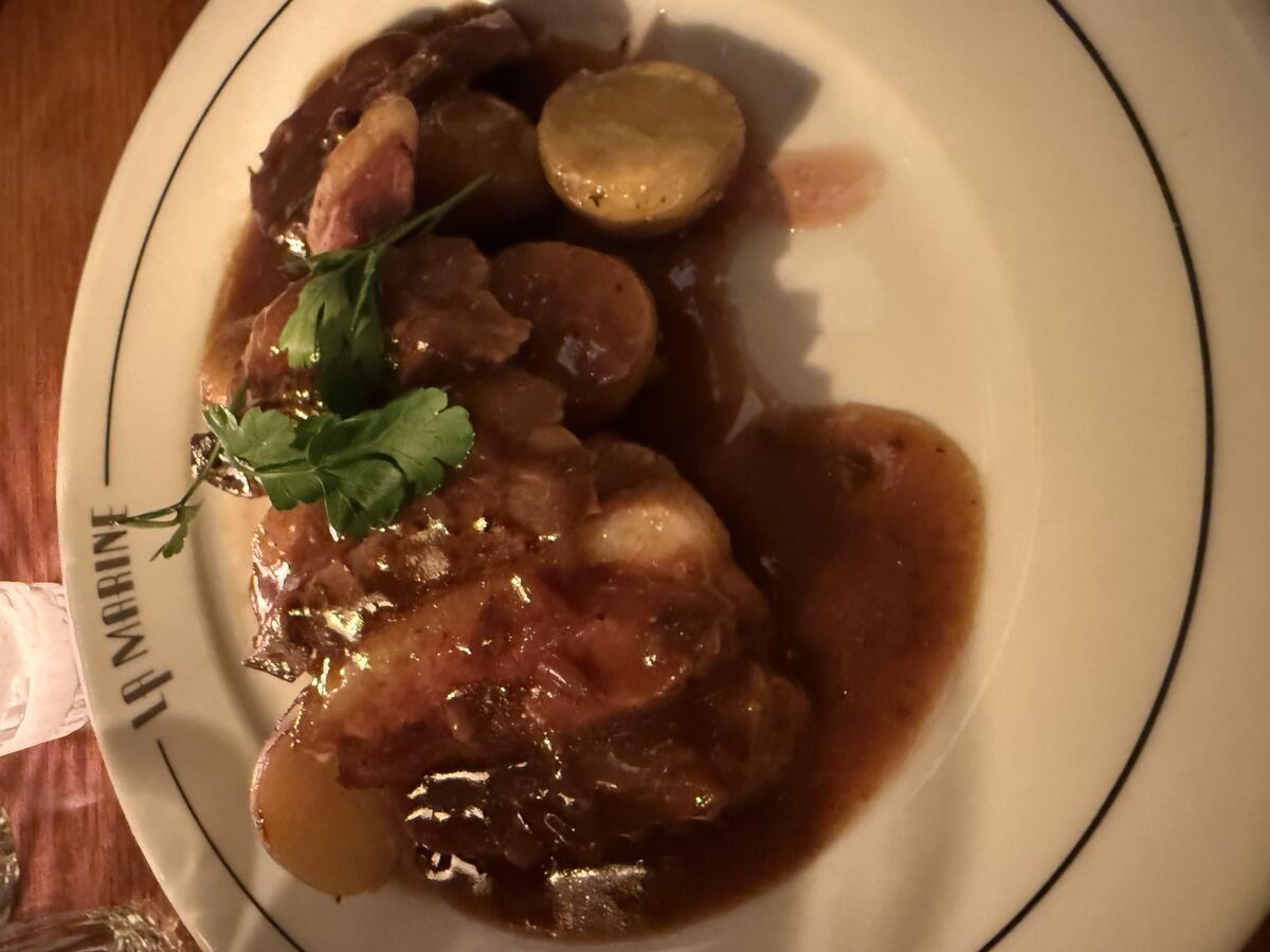 A white plate with a black rim holds a generous serving of sliced roasted meat and small potatoes, all covered in rich brown gravy and garnished with fresh parsley. The name "LA MARINE" is printed on the rim.