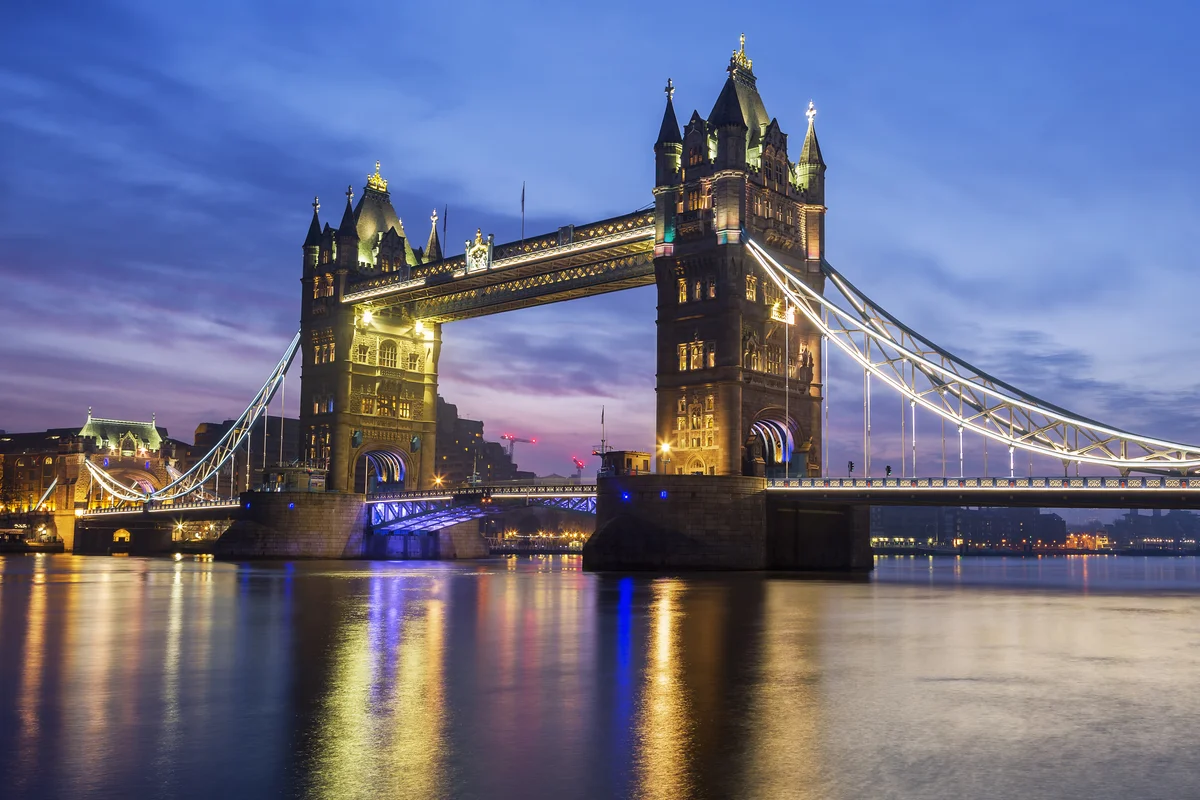 Tower Bridge illuminated at dusk, with glowing lights reflecting across the calm river and a blue‑purple sky creating a dramatic backdrop over the historic structure.