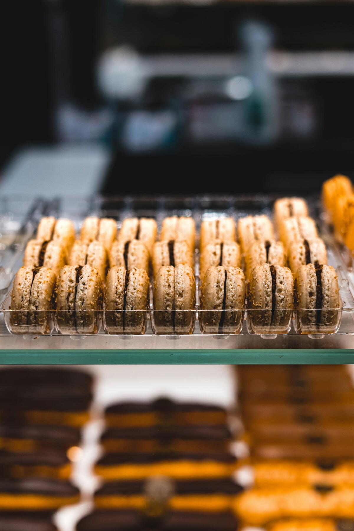 A neatly arranged clear plastic tray filled with light-colored, sandwich-style macarons containing a dark brown filling, displayed in a bakery case.