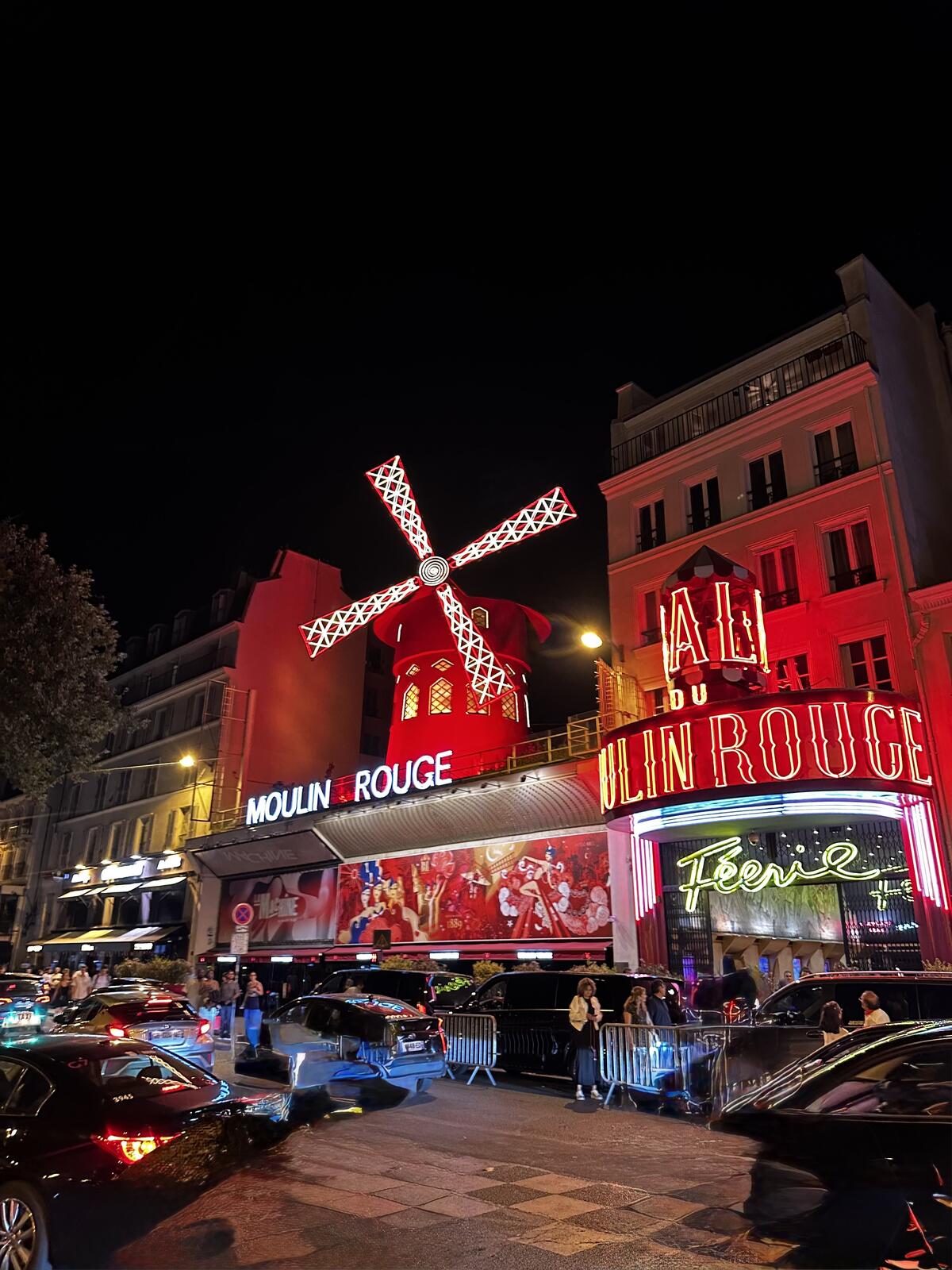 The iconic Moulin Rouge cabaret in Paris, with its illuminated red windmill and "FÉERIE" sign, bustling with people and traffic on a dark night.