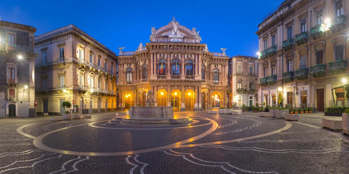 Illuminated historic opera house facing an empty patterned square at dusk.