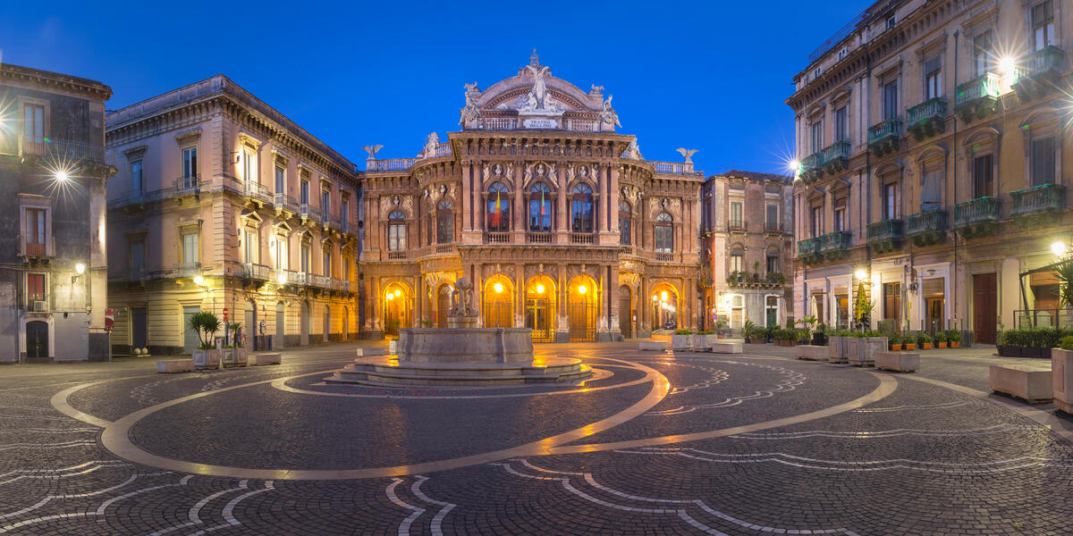 Illuminated historic opera house facing an empty patterned square at dusk.